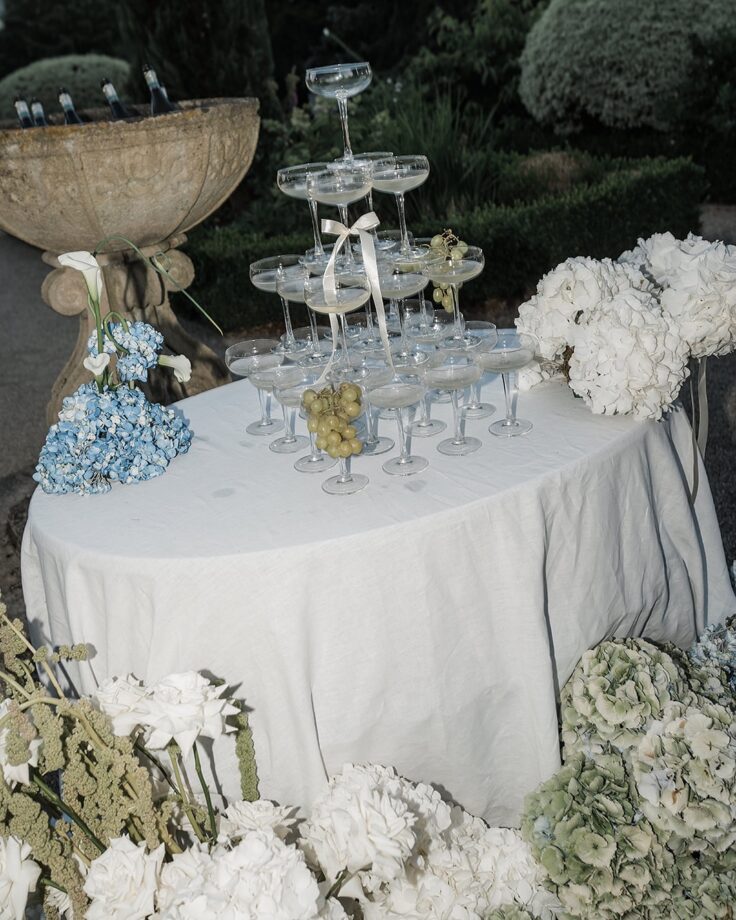 A champagne tower with coupe glasses elegantly stacked on a linen-draped table, adorned with white and blue hydrangeas, fresh green grapes, and a ribbon-tied glass. Website booking clients.